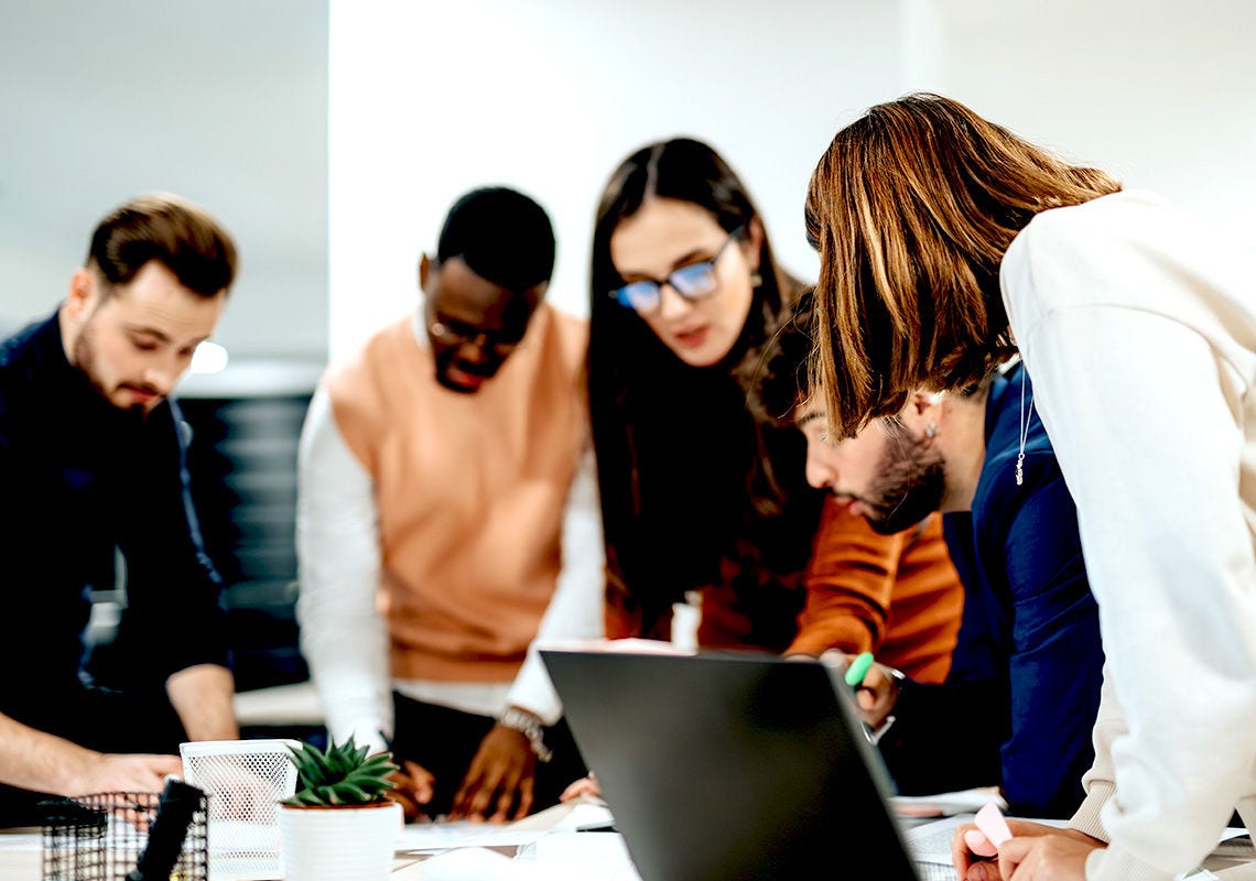 Five people standing around a table, each writing notes or observing. A laptop computer and assorted office supplies are also on the table.