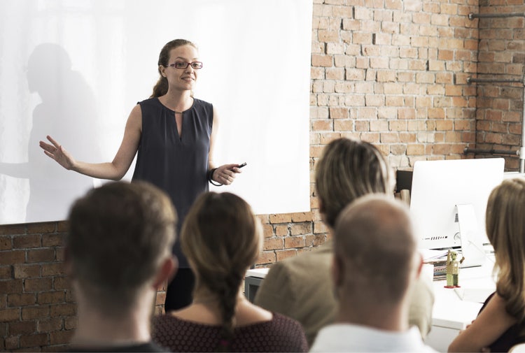 A woman wearing glasses and a dark gray sleeveless shirt speaks in front of a white board while as a group of people listen.