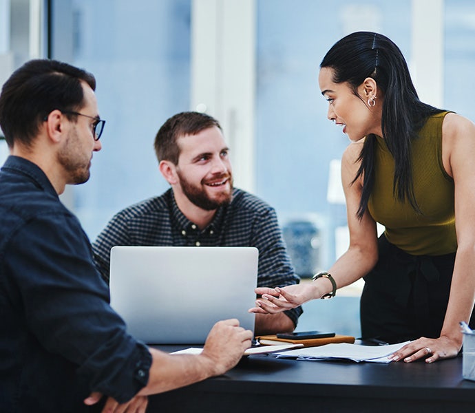 Three people in a shared office space talk at a table. One person is standing and two people are sitting. On the table is a laptop computer and work supplies.