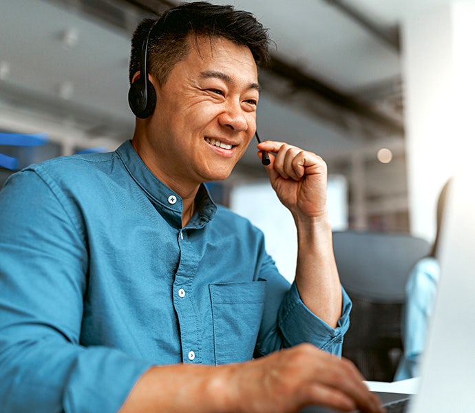 A smiling man in a blue shirt wearing a headset sitting at a desk and working on a computer.