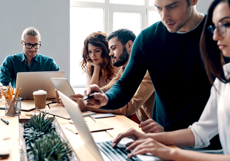 Six people work at a shared desk, each focused on writing in a notebook or typing on a computer.