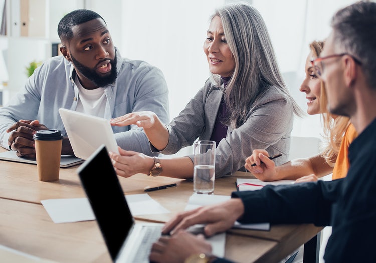 Four people talk while sitting at a table. One woman is holding a tablet. Work supplies and a laptop computer are on the table.
