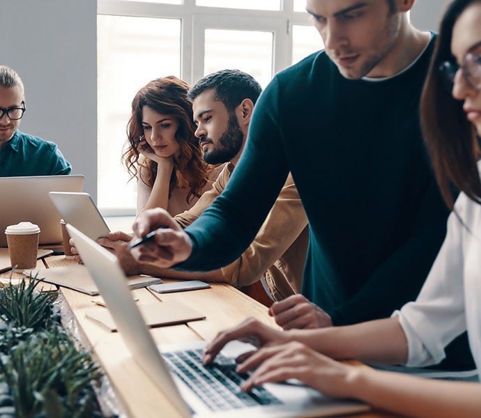 Six people work at a shared desk, each focused on writing in a notebook or typing on a computer.