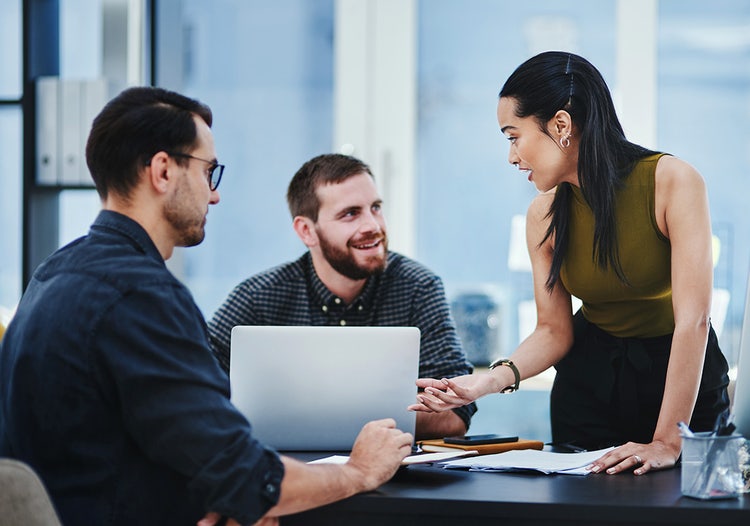Three people in a shared office space talk at a table. One person is standing and two people are sitting. On the table is a laptop computer and work supplies.