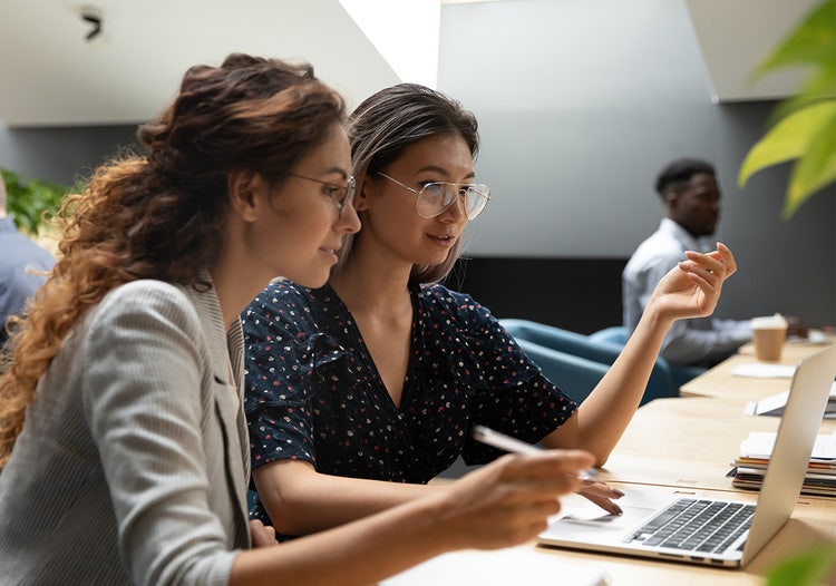 Two women sitting side-by-side at a desk in a shared space, both looking at the same laptop computer screen.