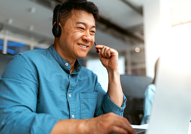 A smiling man in a blue shirt wearing a headset sitting at a desk and working on a computer.