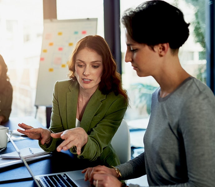 Two women collaborate in front of a laptop computer at a table in a shared office space. In the background, another woman is focused on writing in a notebook.