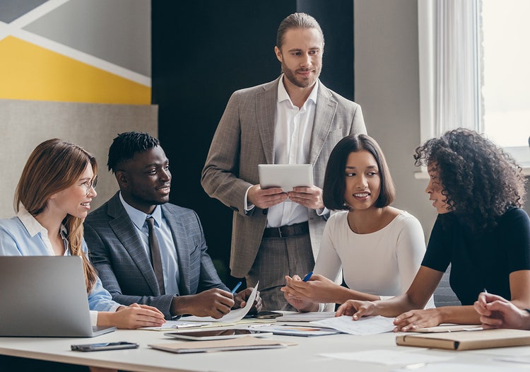 Six people in professional clothing gathering around a table in a conference room. One man is standing in the background while the other five sit.